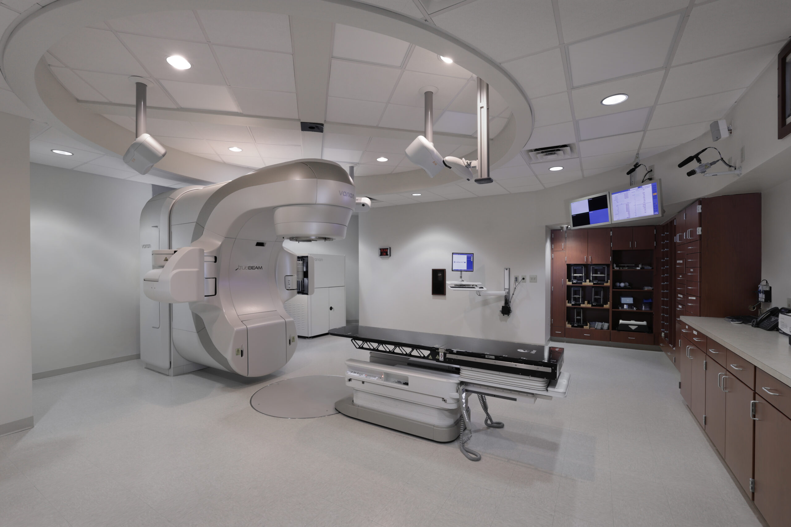 The image depicts a modern medical treatment room designed for radiation therapy, featuring a Varian TrueBeam linear accelerator. The room is clean and well-lit with a neutral color palette, highlighting advanced equipment. Cabinets and storage areas are neatly organized along the walls, and monitors are mounted for operational use. The ceiling has a recessed design with integrated lighting and equipment mounts.