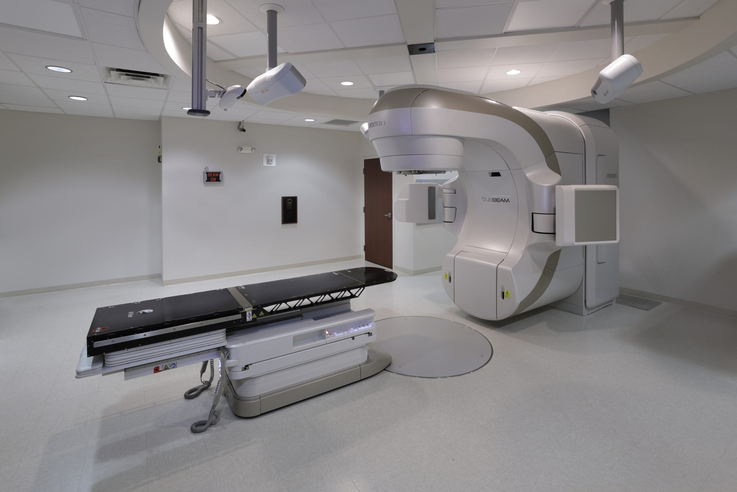 The image shows a clean, modern treatment room with a Varian TrueBeam linear accelerator and a treatment table. The design is neutral and organized, with ceiling-mounted components and a "Beam On" sign on the wall.